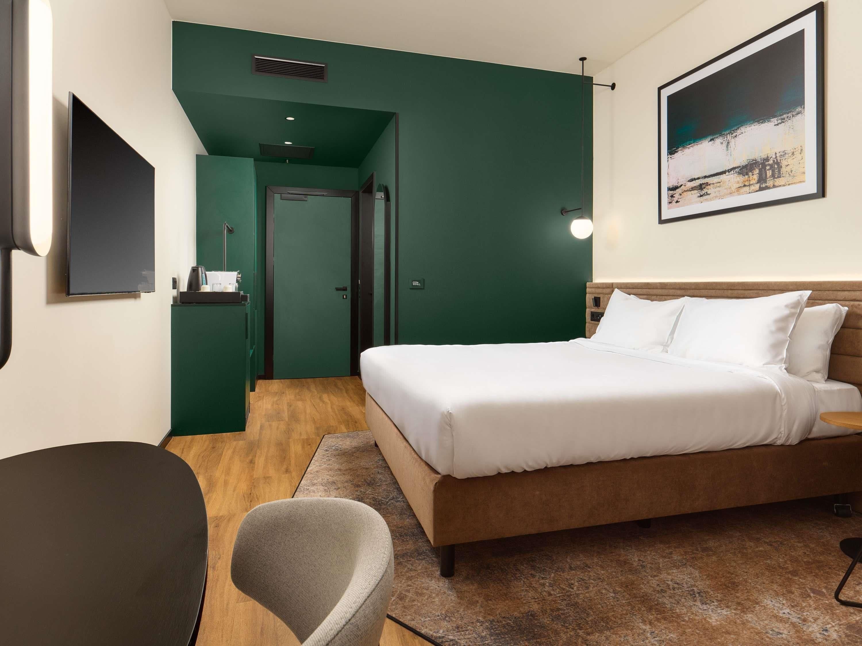 A hotel room with a bed, a chair, a desk, and a painting on the wall.