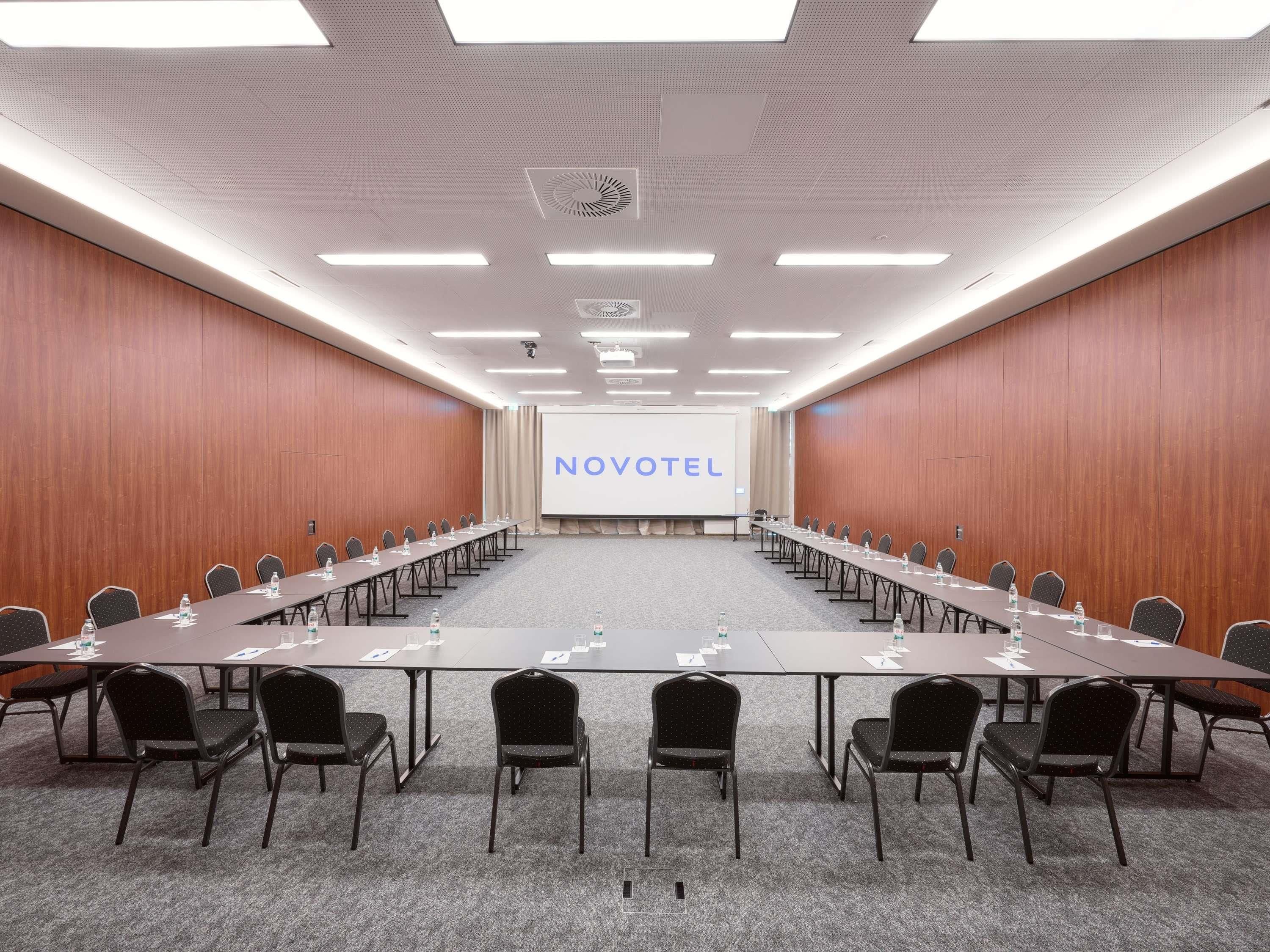 A conference room with a long table, chairs, a projector screen, and a 'NOVOTEL' sign.