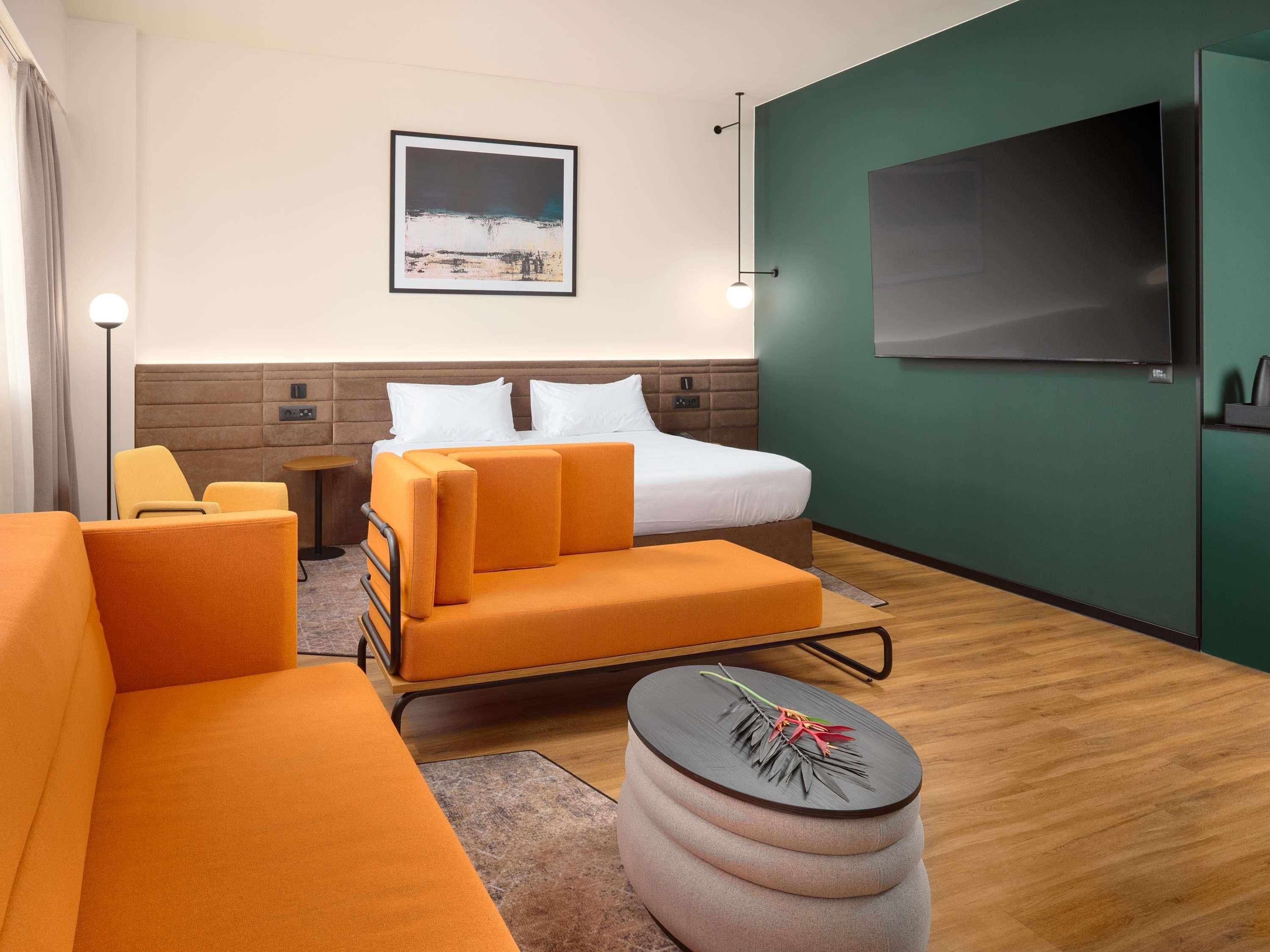 A hotel room with a bed, orange sofa, round ottoman, and a TV on the wall.
