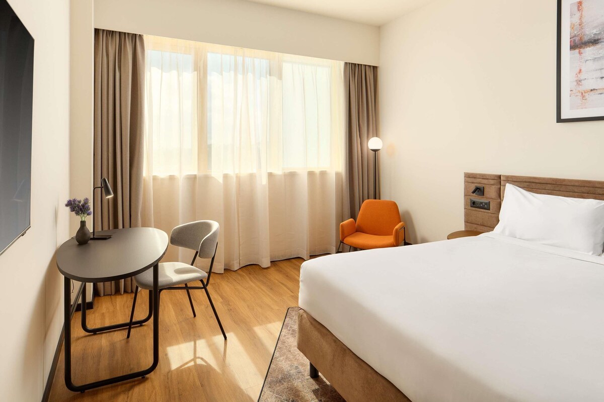 A hotel room with a bed, a desk with a chair, a floor lamp, and a window with curtains.