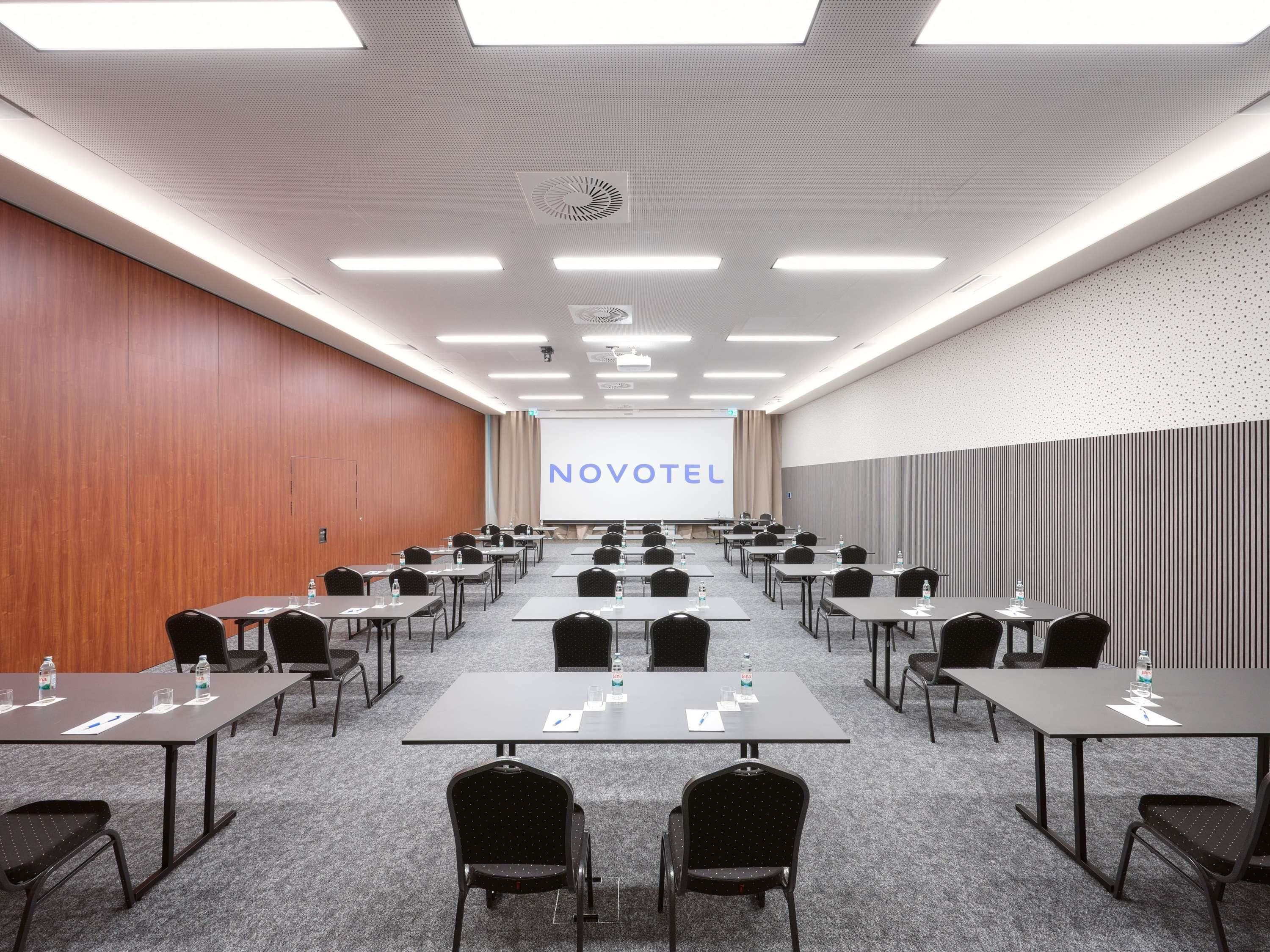 A conference room with multiple tables and chairs, a projector screen displaying 'NOVOTEL', and a ceiling with recessed lighting.