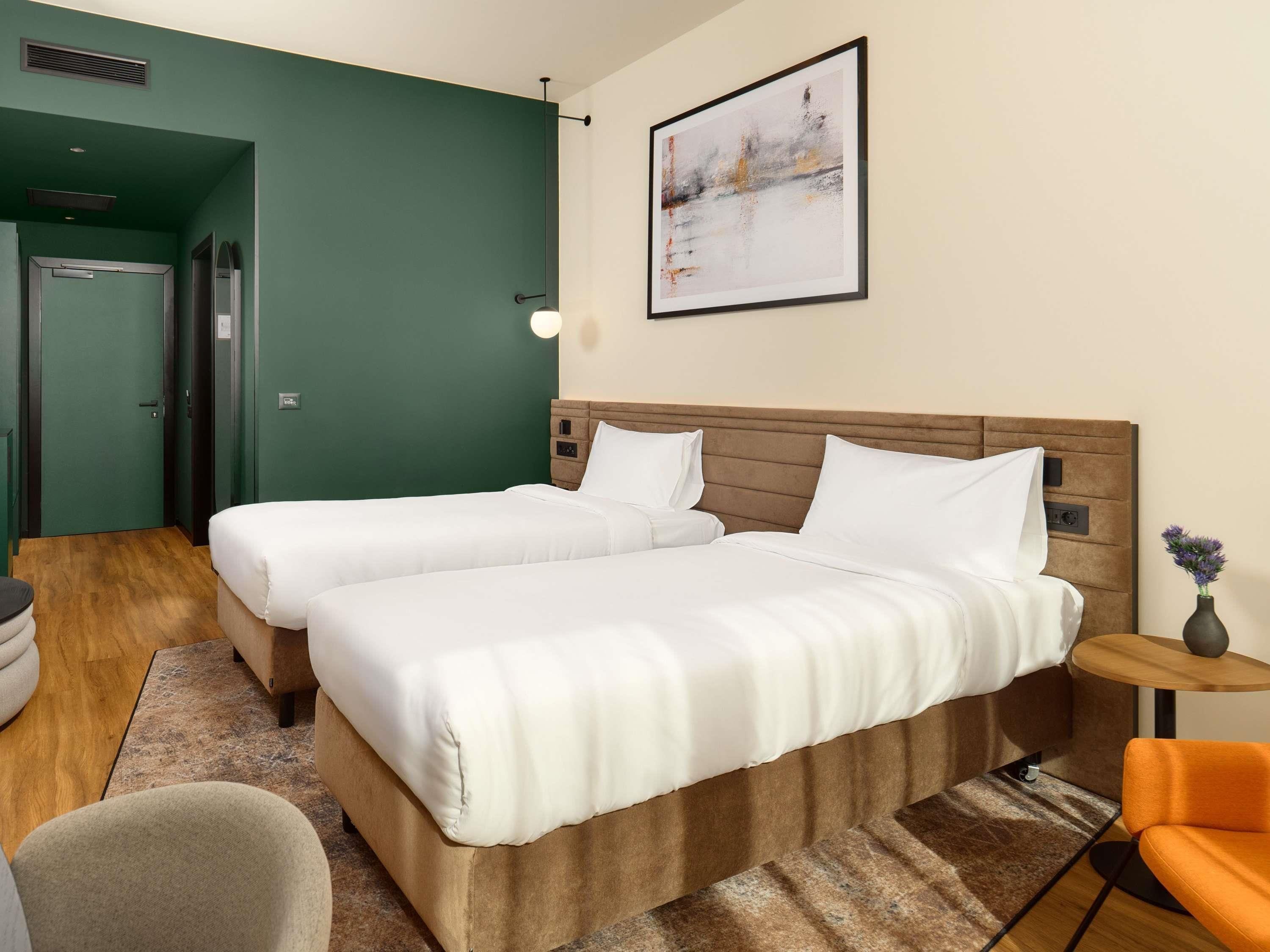 A hotel room with two beds, a wooden headboard, a small round table with a vase, and a painting on the wall.