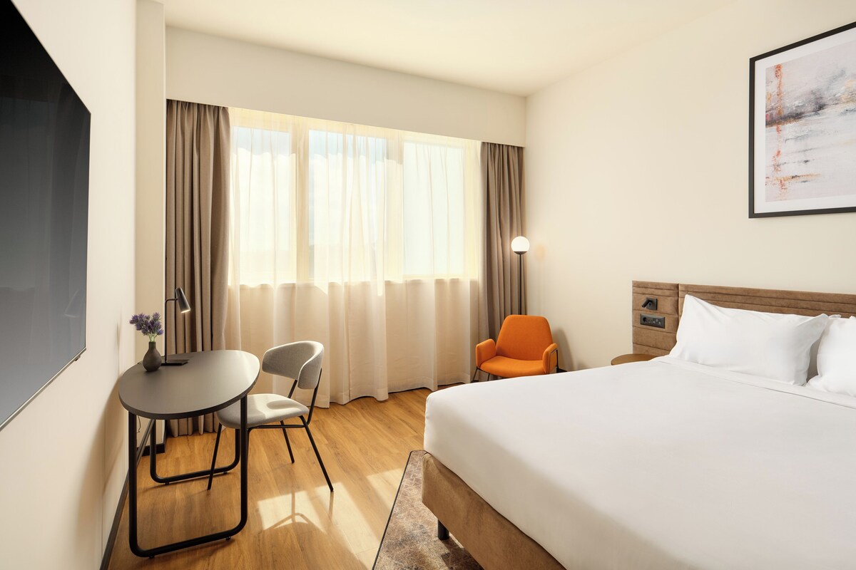 A hotel room with a bed, a desk with a chair, a lamp, and a window with curtains.