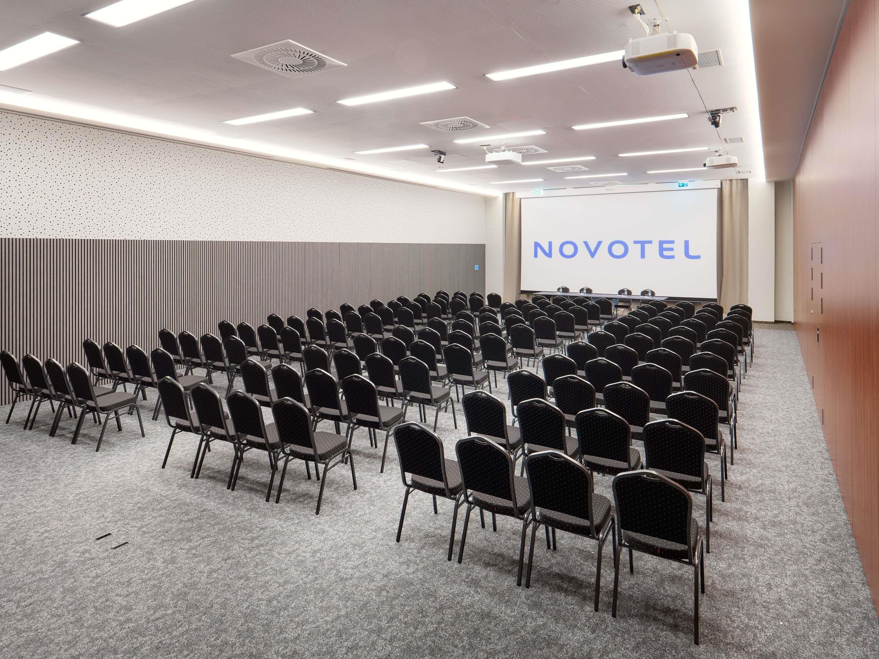 A conference room with rows of chairs, a projector screen, and a 'NOVOTEL' sign.