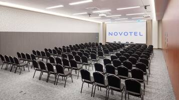 A conference room with rows of chairs, a projector screen, and a 'NOVOTEL' sign.