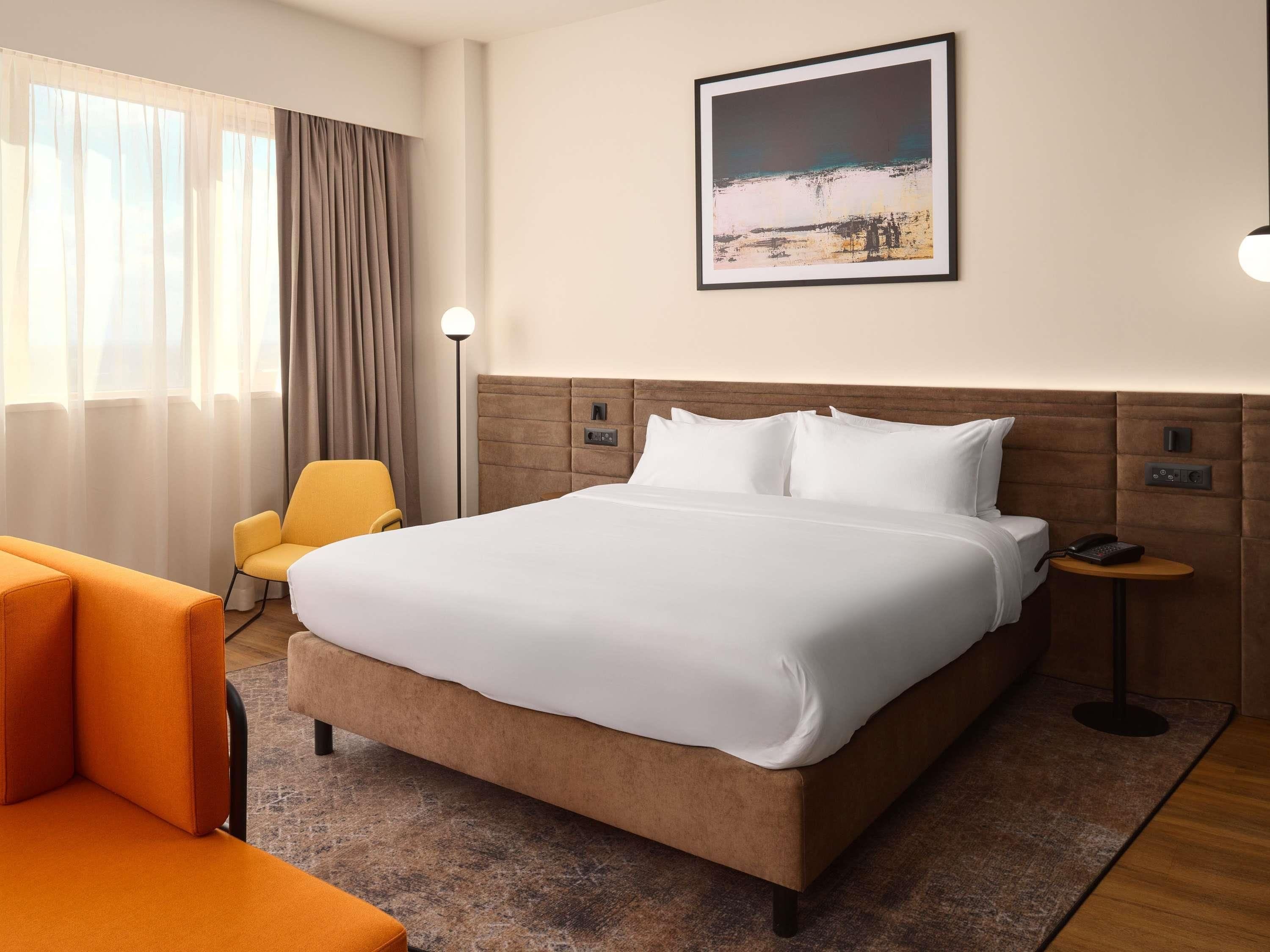 A hotel room with a large bed, a chair, a bedside table with a phone, and a painting on the wall.