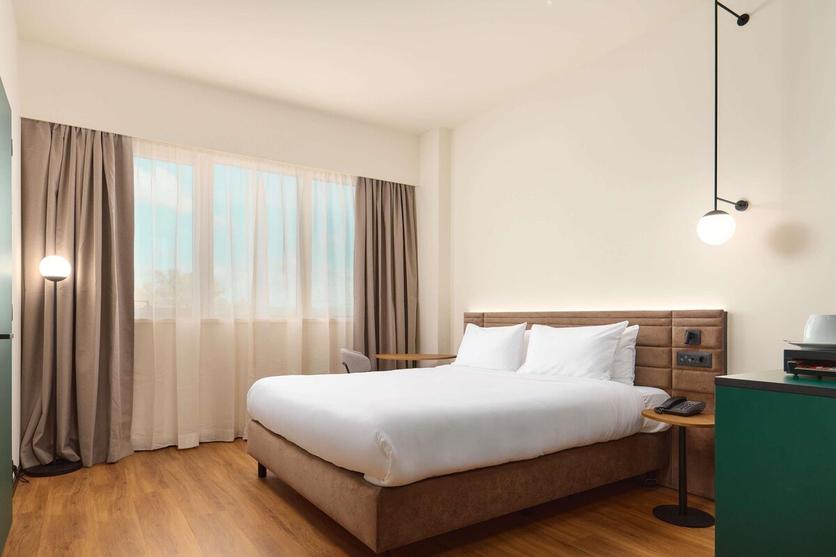 A hotel room with a bed, curtains, a bedside table with a lamp, and a nightstand with a phone.
