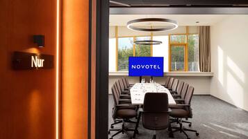 Ein Konferenzraum mit einem langen Tisch, Stühlen und einem Fernsehbildschirm, der 'NOVOTEL' anzeigt.