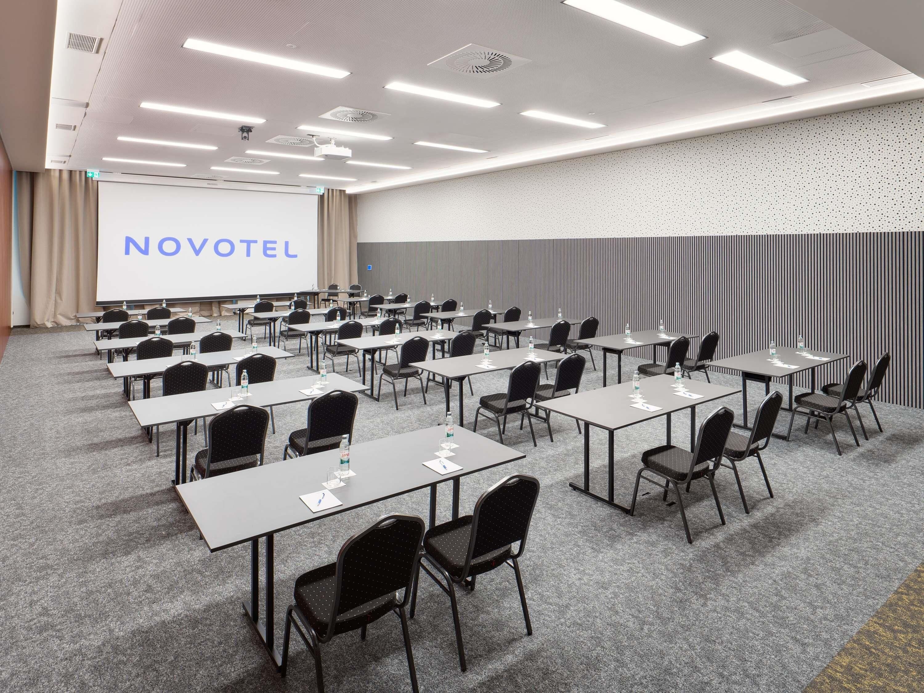 A conference room with rows of tables and chairs, a projection screen, and a 'NOVOTEL' sign.