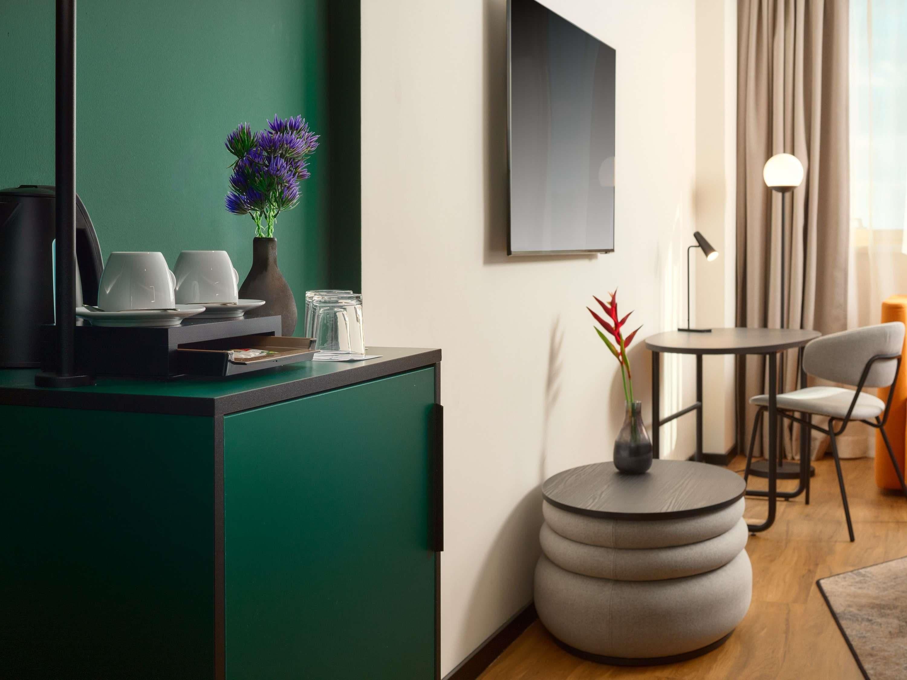 Una habitación de hotel moderna con una pared verde de acento, una estación de café, una mesa redonda con sillas y un ventanal con cortinas.