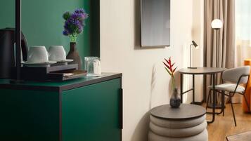 Una habitación de hotel moderna con una pared verde de acento, una estación de café, una mesa redonda con sillas y un ventanal con cortinas.