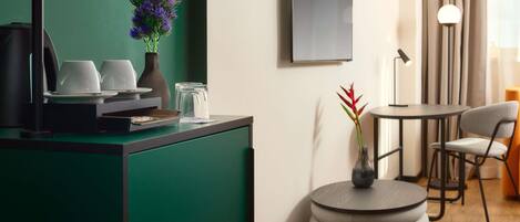 Una habitación de hotel moderna con una pared verde de acento, una estación de café, una mesa redonda con sillas y un ventanal con cortinas.