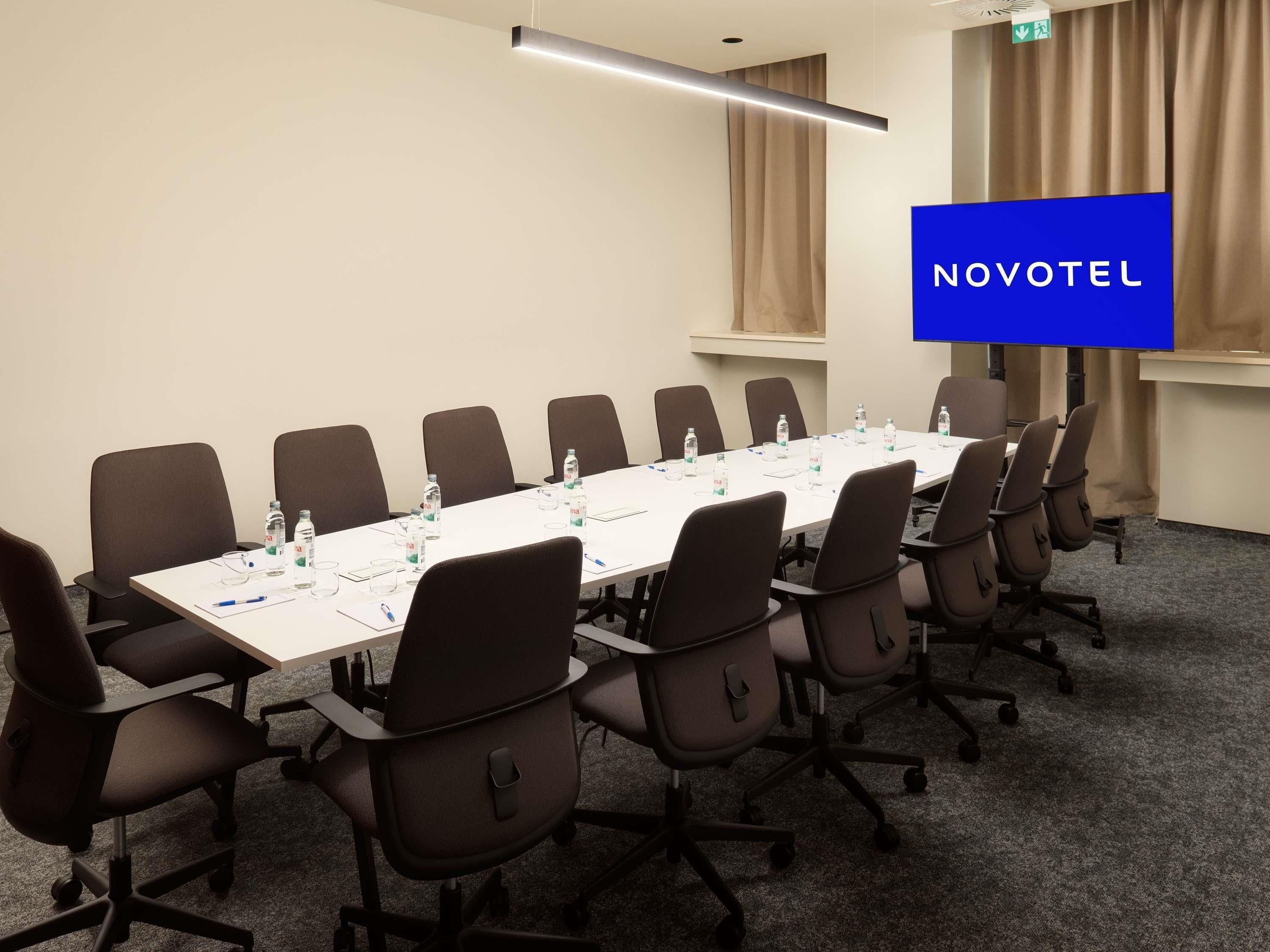 Et mødelokale med et langt bord, stole, vandflasker og et blåt skilt med 'NOVOTEL' på væggen.