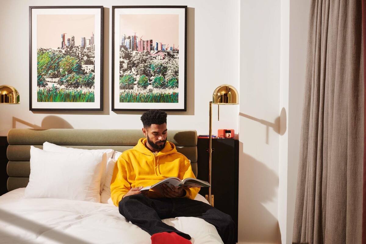 A person sitting on a bed in a hotel room, reading a book. There are two framed pictures on the wall, a floor lamp, and a bedside table with a radio.