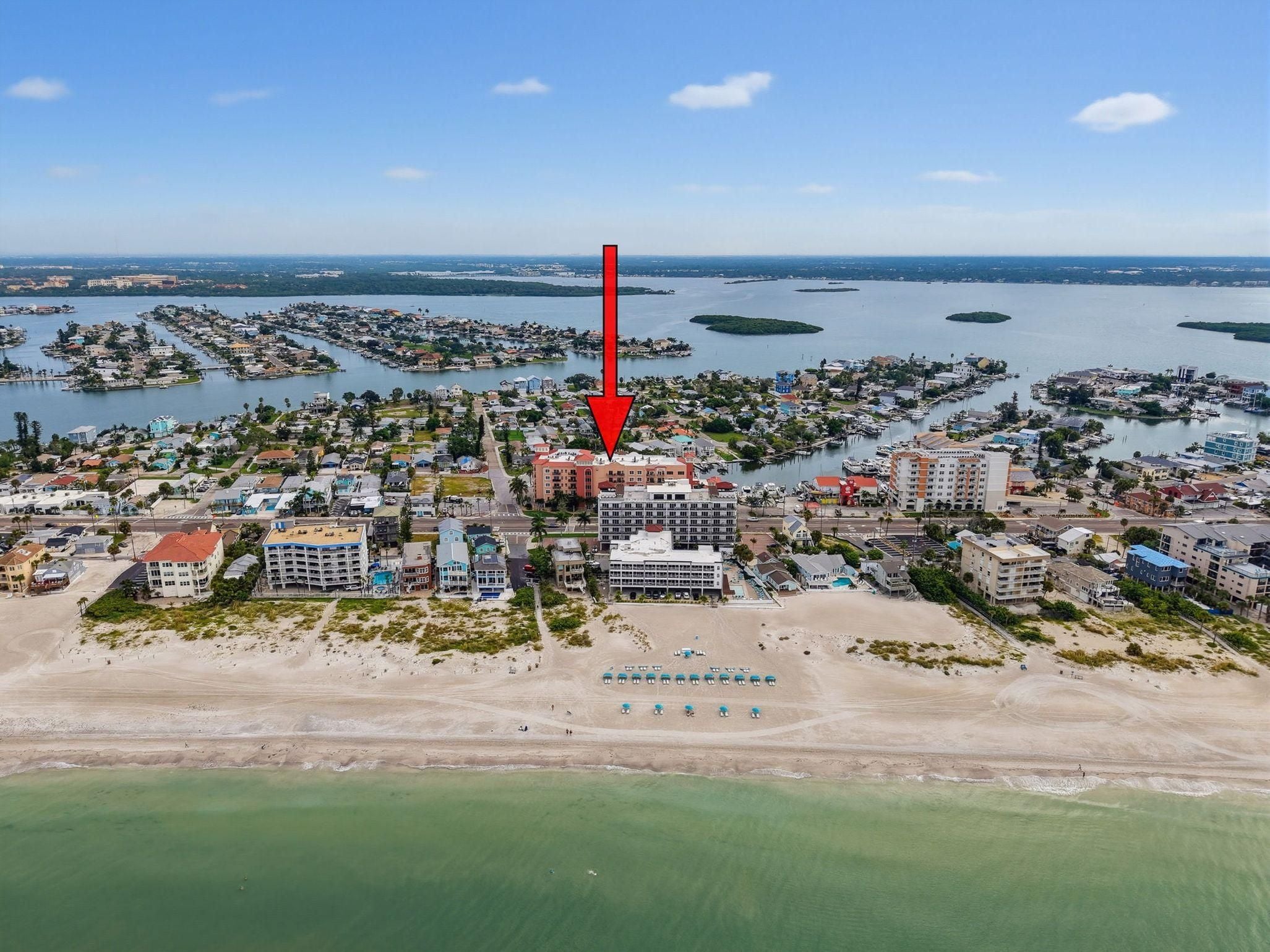 An aerial view of a coastal town with a prominent hotel marked by a red arrow, surrounded by residential areas and water bodies.