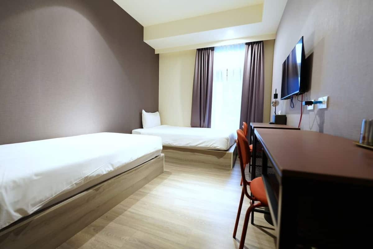 Habitación de hotel con dos camas individuales, un escritorio con silla, un televisor de pantalla plana montado en la pared y una ventana con cortinas.