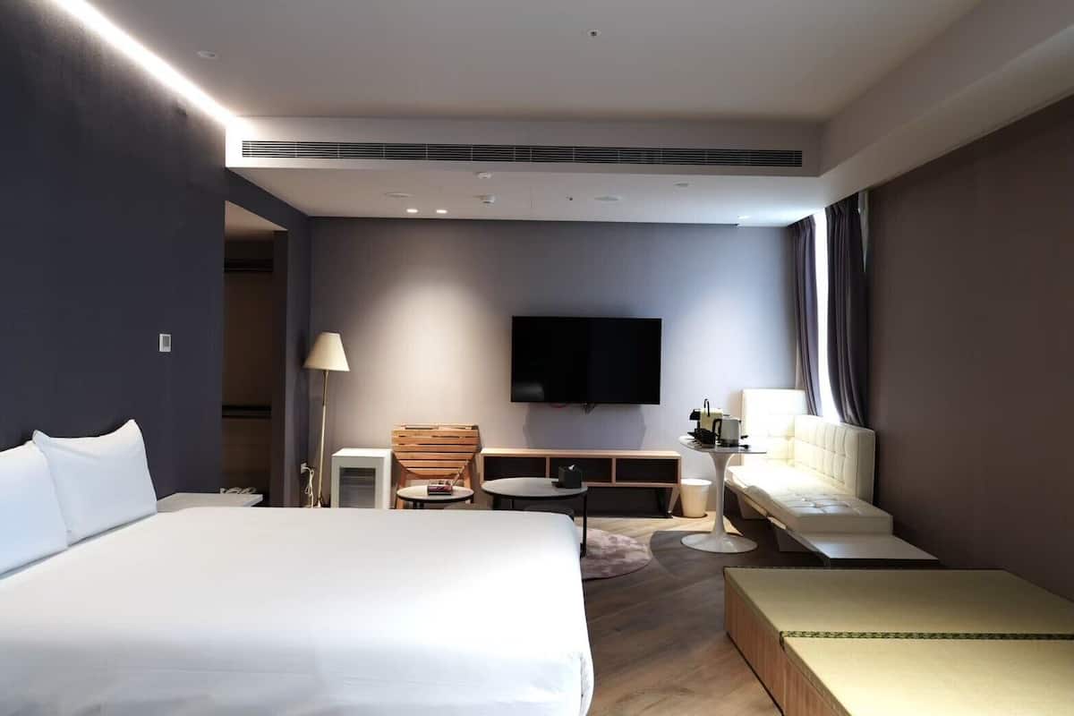 Una habitación de hotel moderna con una cama grande, un televisor de pantalla plana, un área de estar con una mesa y sillas, y un sofá.