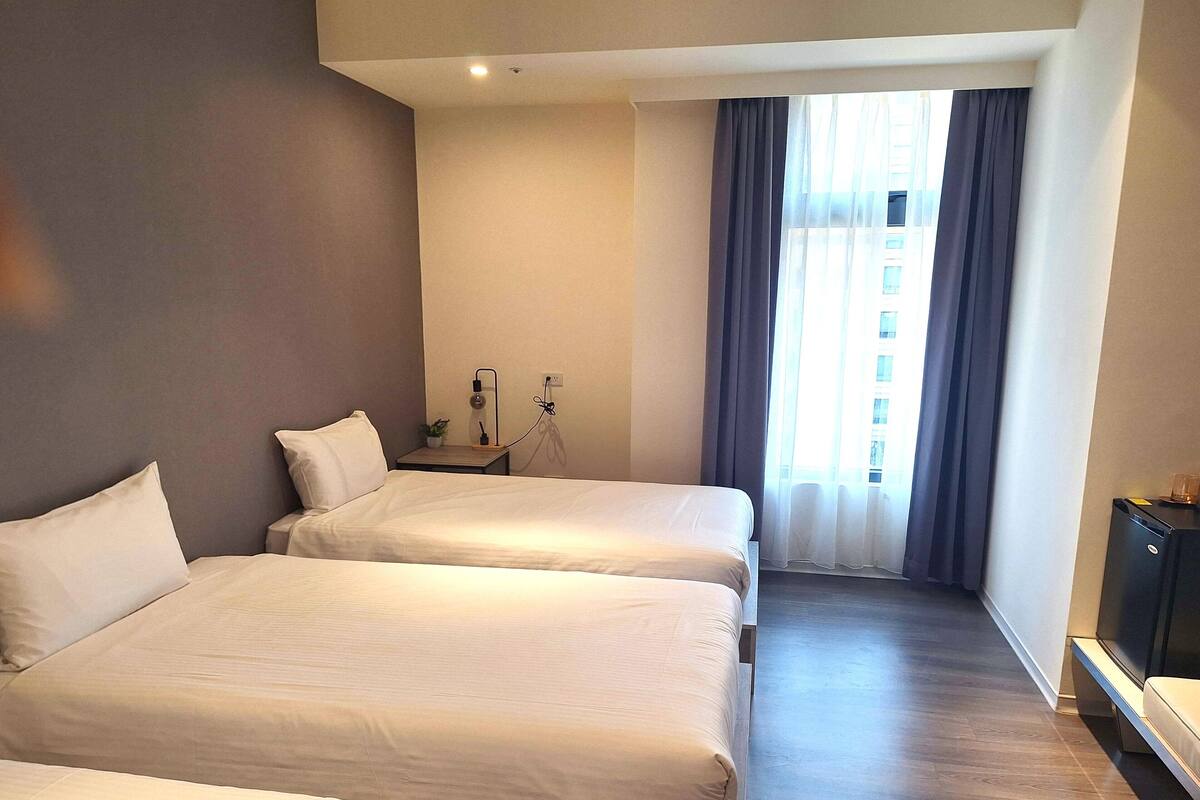 Habitación de hotel con dos camas, una mesita con una planta, un escritorio con una computadora y una ventana con cortinas.