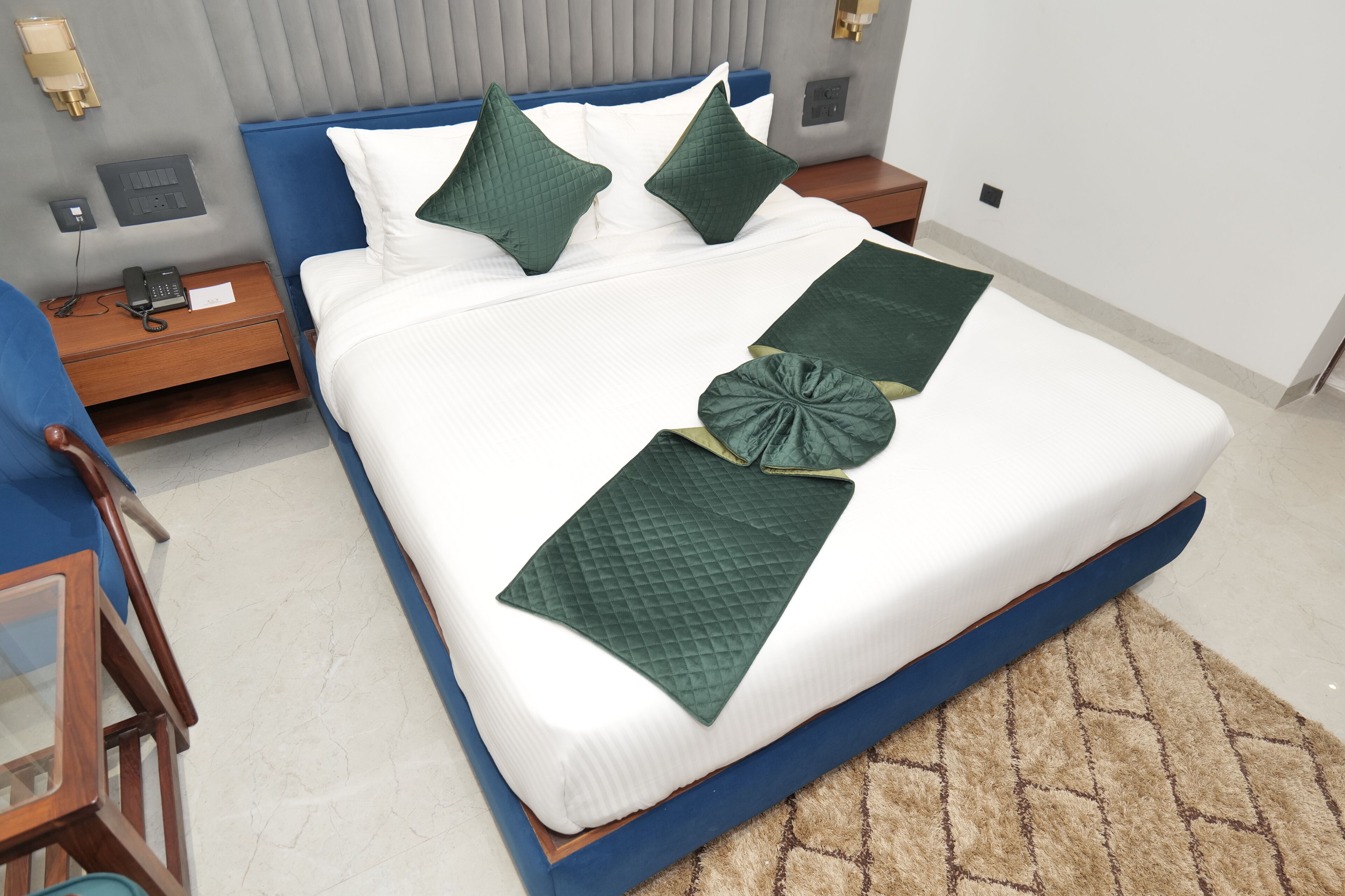 A neatly made bed with green and white bedding, a bedside table with a phone, and a nightstand with a lamp.