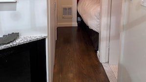 Ein schmaler Flur mit Holzboden, eine weiße Tür rechts und ein Bett mit weißer Bettwäsche im angrenzenden Zimmer.