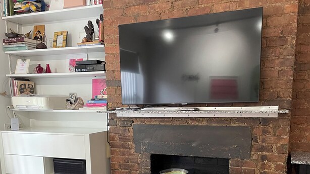 Ein Zimmer mit Backsteinwand, einem flachbildfernseher über einem Kamin, einer weißen Schrank mit Regalen und einem Esstisch mit zwei Stühlen.