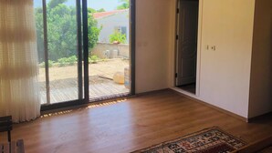 A room with a wooden floor, a sliding glass door leading to a garden, a patterned rug, and a ceiling light fixture. - Incekum Pansiyon (Maziköy)
