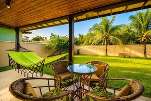 A covered patio with wicker furniture, a hammock, and a view of a lush garden. - Bz98 Geribá casa 4 suítes e área gramada (Buzios)