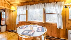 A wooden cabin interior with a round dining table, chairs, a window with curtains, and wall-mounted lamps.