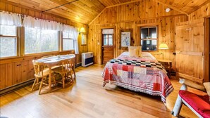 A wooden cabin interior with a dining table, chairs, a bed with a quilt, and a window with curtains.