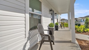A covered porch with two chairs and potted plants, a clear sky, and neighboring houses.