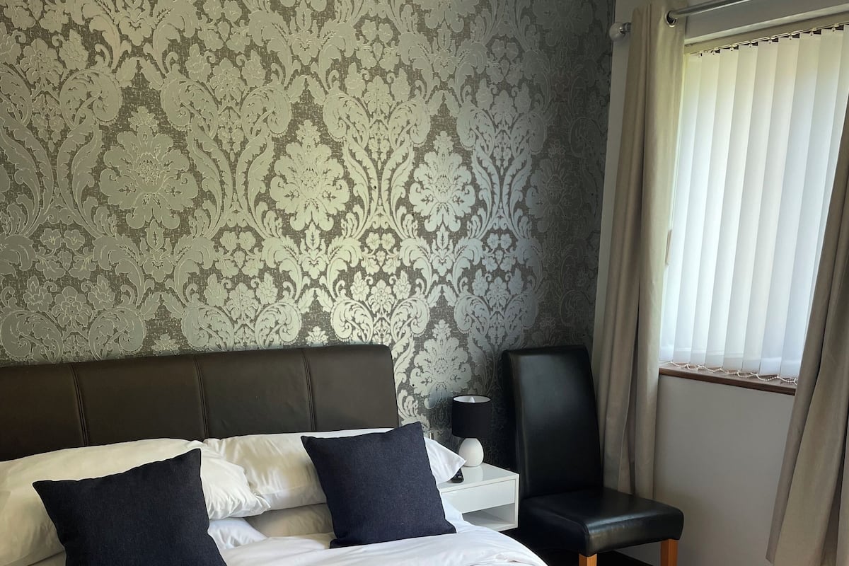 Una cama bien hecha con sábanas blancas, dos almohadas oscuras y una toalla enrollada. Hay una mesita de noche con una lámpara y un control remoto. La habitación tiene papel tapiz estampado y una ventana con cortinas.