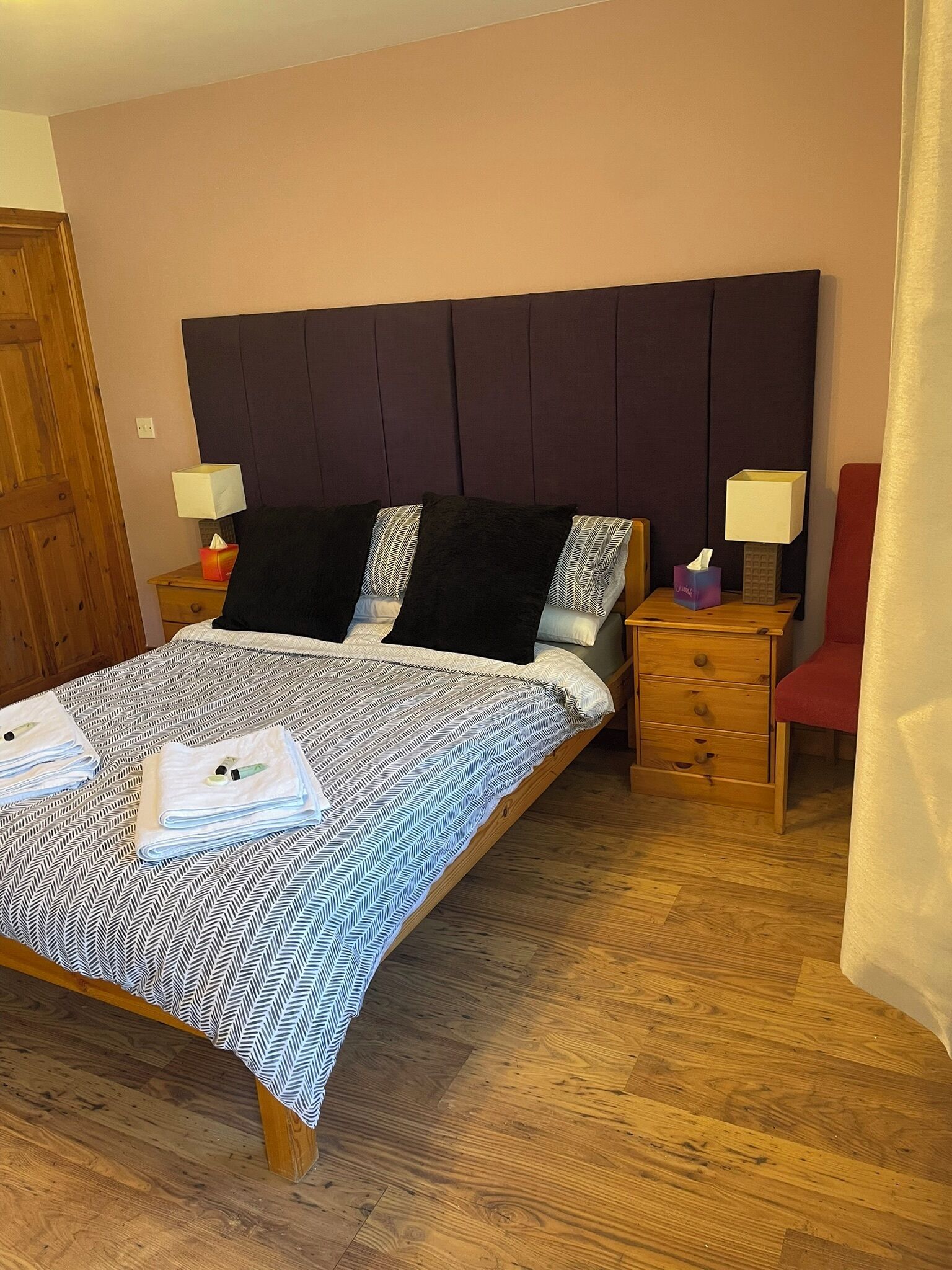 A neatly made bed with a patterned bedspread, a wooden headboard, a bedside table with a lamp, a red chair, and a wooden wardrobe.