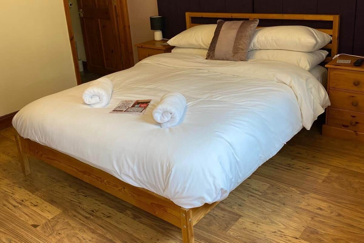 Una cama bien hecha con sábanas blancas y dos toallas enrolladas, cabecera de madera, mesita de noche con lámpara y suelo de madera.