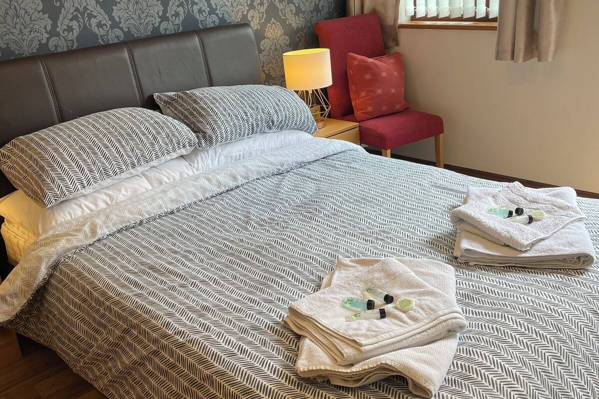 Una cama bien hecha con ropa de cama estampada, una silla roja, una lámpara y una ventana con cortinas.