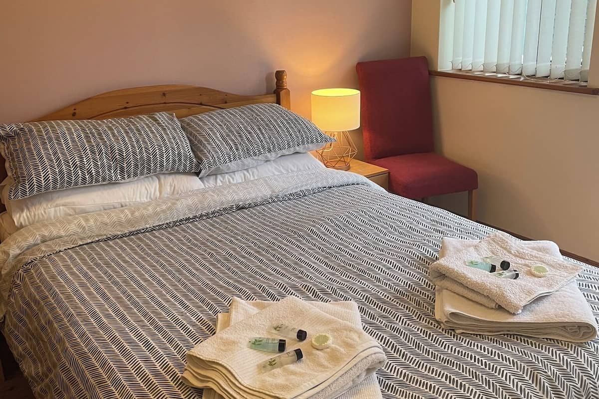 Una cama bien hecha con ropa de cama estampada, una lámpara de noche, una silla roja y una ventana con persianas.