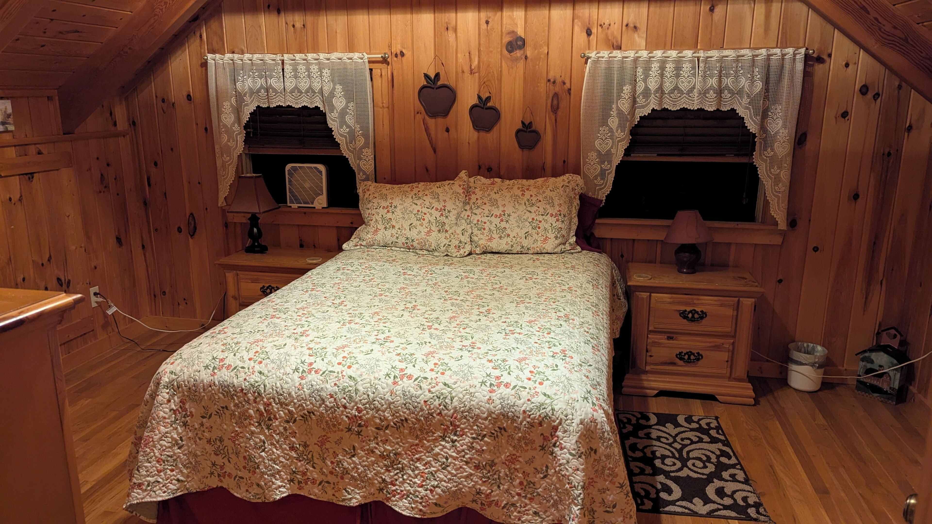 A cozy bedroom with a floral bedspread, wooden walls, and a small bedside table with a lamp.
