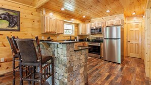 A rustic kitchen with wooden cabinets, a stone island, a stainless steel refrigerator, and a bar stool. - Brand New! Hickory Wind: Charming 2 Bedroom Cabin in Whittier (Whittier)