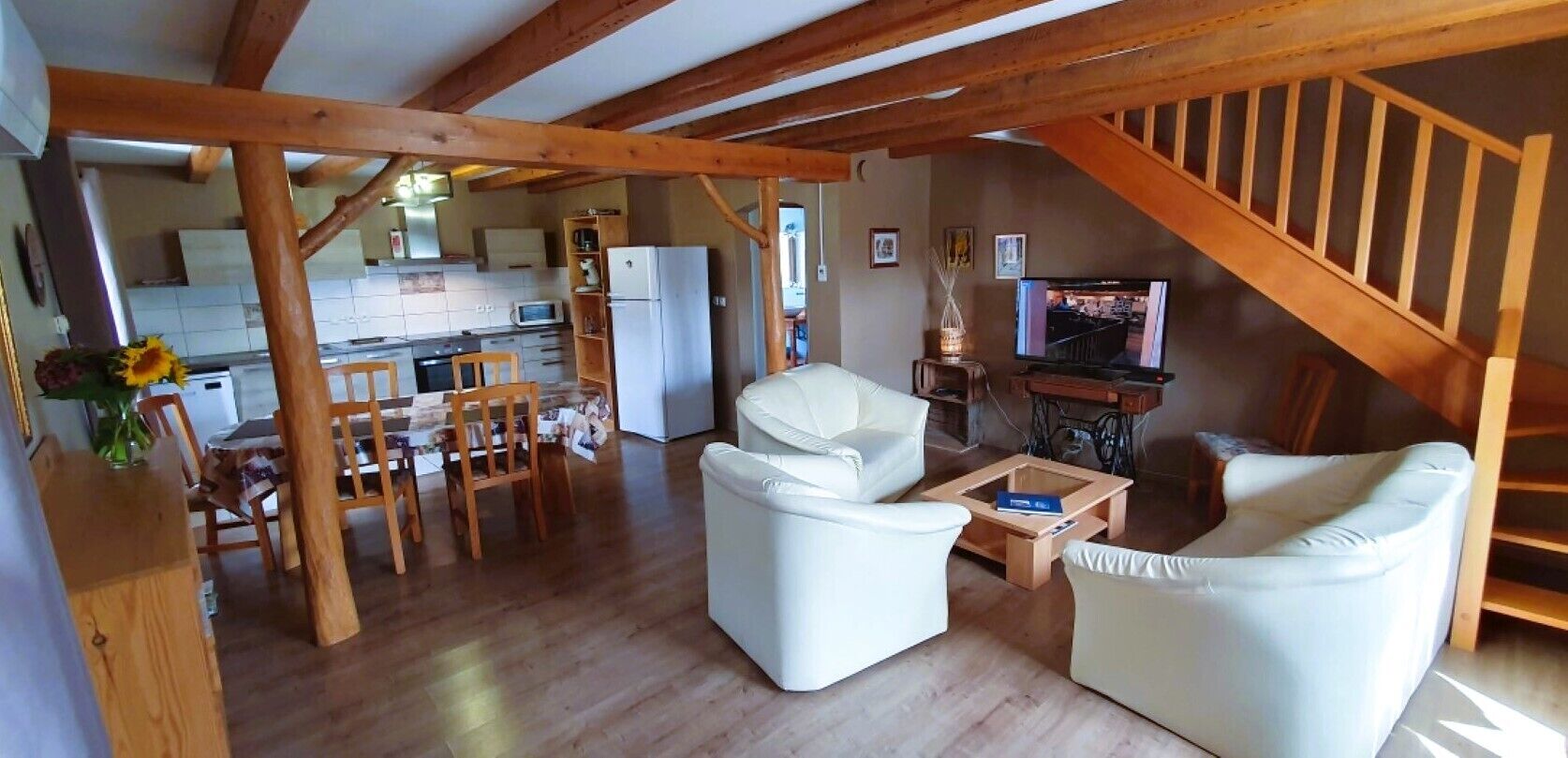 A spacious living area with a wooden staircase, a dining table with chairs, a sofa, and a television.