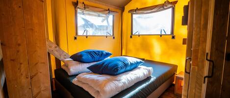 A single bed with blue and white bedding, two windows with curtains, and wooden walls and floor.