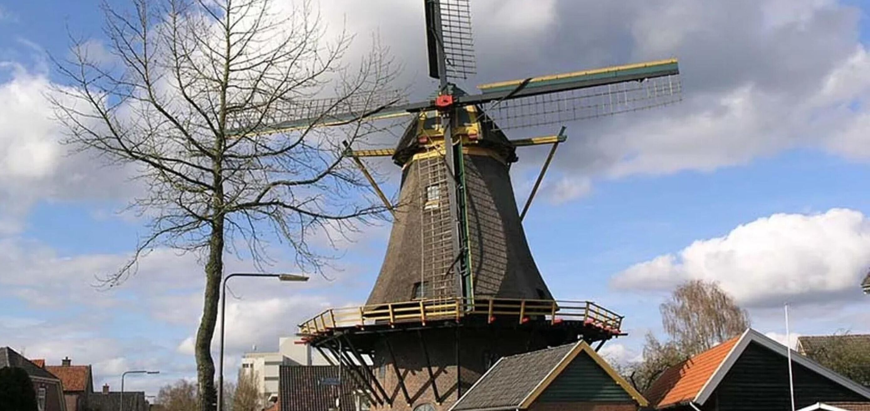 A traditional windmill with a conical roof and a large sail, surrounded by residential buildings and a leafless tree.