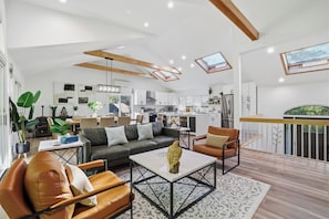 A modern living room with a sofa, coffee table, and chairs, a kitchen in the background, and skylights.