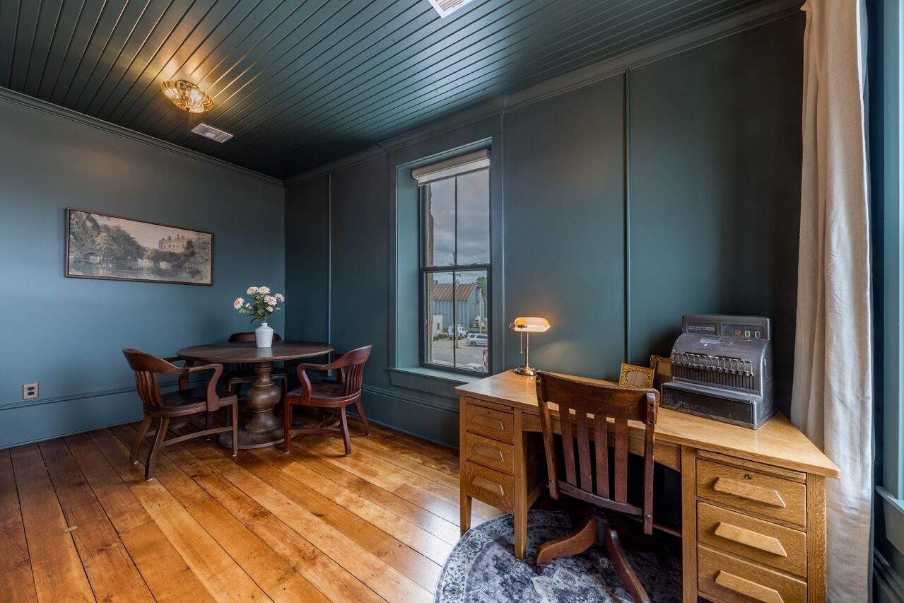 A room with a wooden dining table and chairs, a desk with drawers, a vintage radio, and a window with a view of the outside.