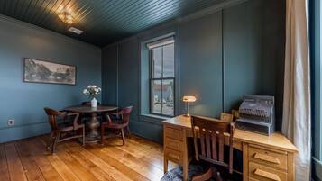 A room with a wooden dining table and chairs, a desk with drawers, a vintage radio, and a window with a view of the outside.