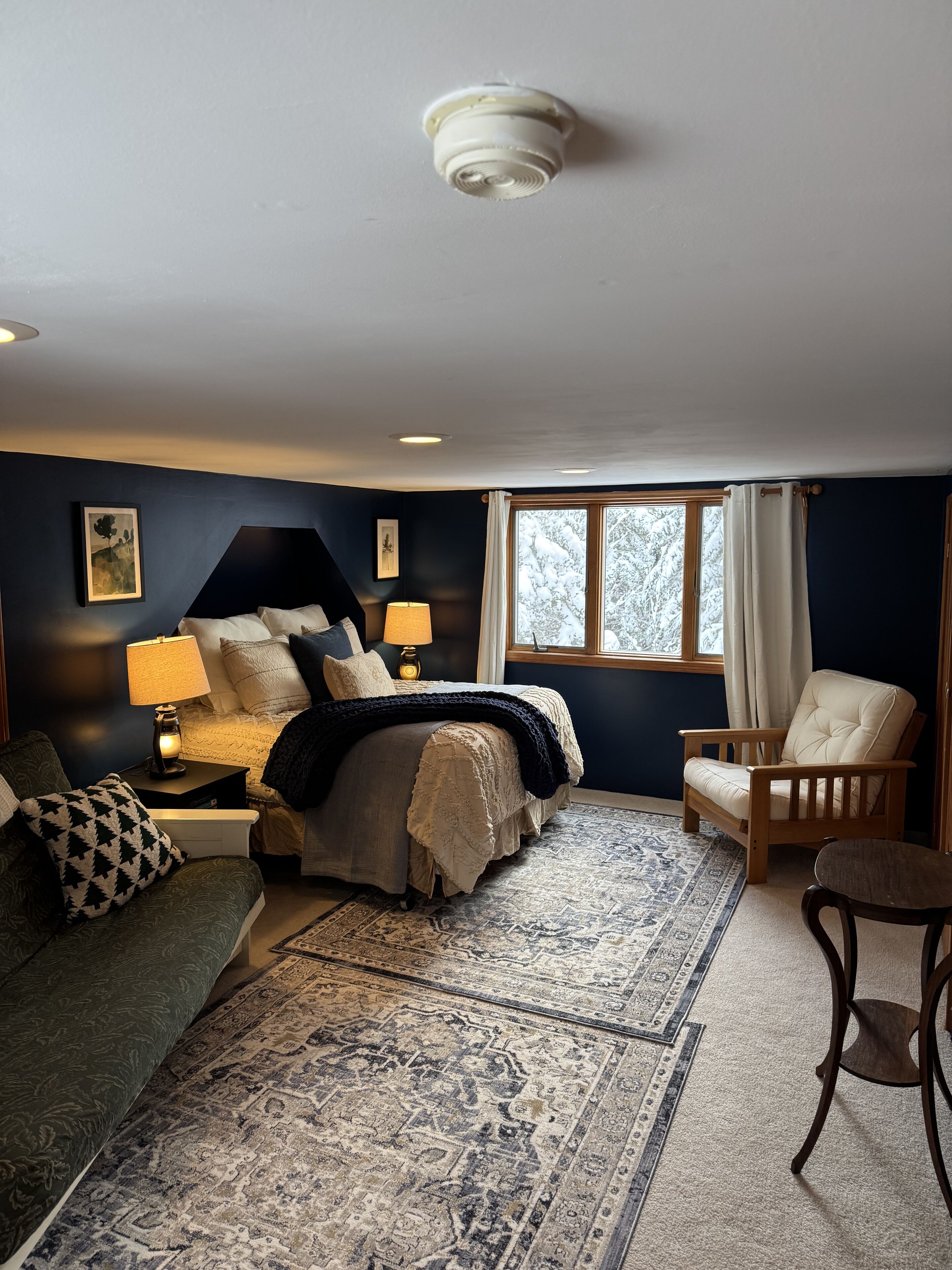 A bedroom with a bed, a chair, a nightstand, a lamp, and a window with curtains.