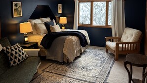 A bedroom with a bed, a chair, a nightstand, a lamp, and a window with curtains.