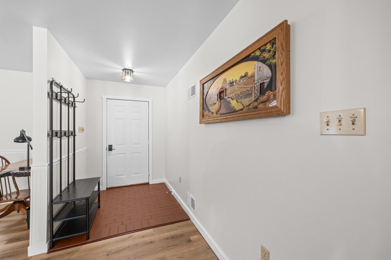 Un couloir avec une porte blanche, un porte-manteau, une table à manger avec des chaises et un tableau encadré accroché au mur.