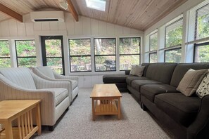 Ein gemütliches Wohnzimmer mit einem Ecksofa, einem Zweisitzer und einem Holztisch. Große Fenster bieten Blick ins Freie.