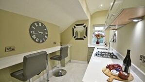 A modern kitchen with a large wall clock, a gas stove, and a wooden cutting board with food items. - Moat House Room 5|Private Bathroom (Rossendale)