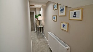 A hallway with a radiator, framed artwork on the walls, and a table with a lamp and flowers. - Moat House Room 5|Private Bathroom (Rossendale)