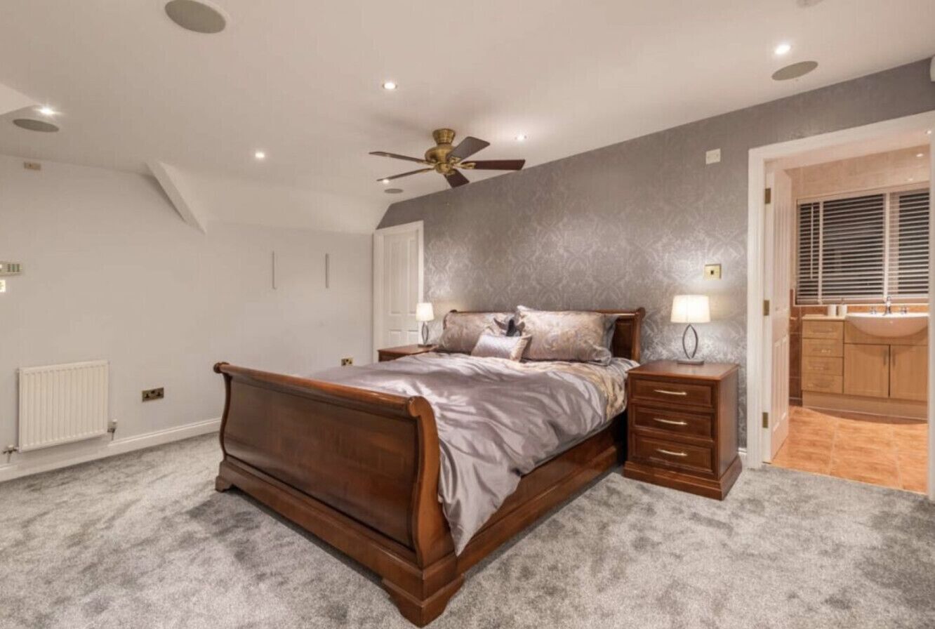 Une chambre à coucher avec un lit en bois, une table de chevet, un ventilateur de plafond et une salle de bain visible à travers une porte ouverte.